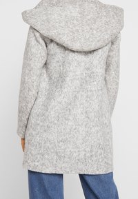 Femme portant un manteau à capuche gris clair texturé, associé à un jean en denim bleu, vue de dos sur un fond blanc uni.