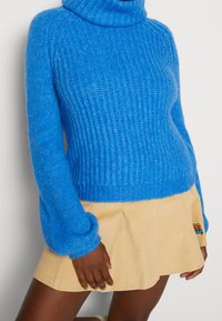 Pull à col roulé côtelé bleu à manches longues associé à une mini-jupe en velours côtelé beige ornée d'un petit écusson brodé.