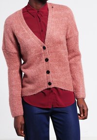 Lyserød strikket cardigan med V-hals, ribbet struktur og sorte knapper. Kombineret med en mørkerød skjorte med knapper under. Ingen synlige mønstre.