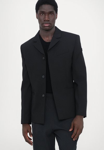 Blazer nero con un taglio moderno, chiusura a un solo bottone, collo a punta e dettagli delle tasche minimalisti. Indossato con un top nero e pantaloni.