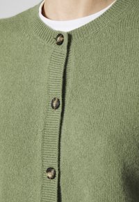 Zelený pletený cardigan s kulatým výstřihem, strukturou tkaniny a kontrastními knoflíky vpředu. Detailní pohled na detaily.