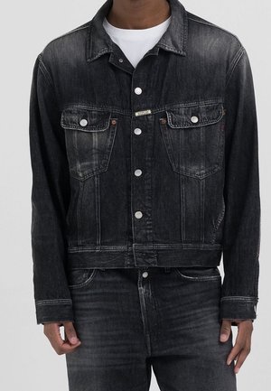 Homme portant une veste en jean noire avec des boutons argentés sur une chemise blanche et un jean noir assorti, debout devant un fond uni.