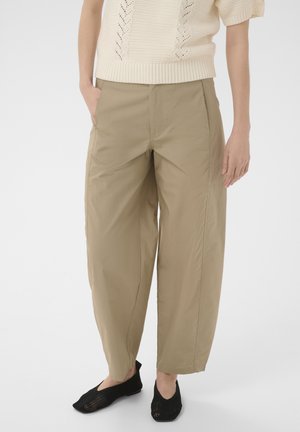 Personne portant un pantalon beige à jambes larges, un pull court en maille crème à manches courtes, et des chaussures slip-on en mesh noir, debout avec une main dans la poche.