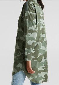 Camisa larga de camuflaje verde con un corte holgado, que presenta un dobladillo en punta, mangas largas y un bolsillo de parche en el pecho. Detalle de tela texturizada en los hombros.