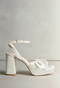Biele platformové sandále s masívnym podpätkom, saténovým povrchom, nastaviteľným remienkom okolo členka a mašľou na prednej časti topánky.