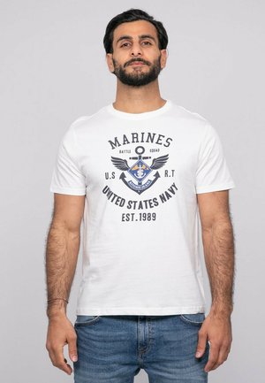 Homme barbu portant un t-shirt blanc avec le texte "Marines United States Navy" et un graphisme d'ancre, associé à un jean bleu, sur un fond gris.