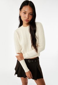 Suéter de punto trenzado color crema, con cuello redondo y mangas largas, combinado con una falda plisada marrón oscuro que cuenta con trabillas y un hebilla.