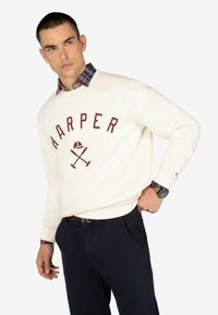 Sudadera blanca con la palabra "HARPER" en letras color marrón, un gráfico de bates cruzados y una gorra. Superpuesta a una camisa de cuadros azul y roja por debajo. Pantalones azul marino.