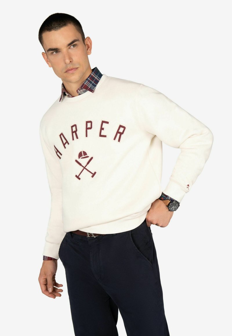 Sudadera blanca con la palabra "HARPER" en letras color marrón, un gráfico de bates cruzados y una gorra. Superpuesta a una camisa de cuadros azul y roja por debajo. Pantalones azul marino.