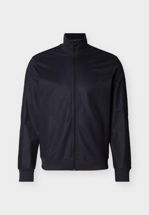 Ναυτικό μπλε τζάκετ με φερμουάρ, φτιαγμένο από απαλό ύφασμα, με ψηλό γιακά, ριμπ μανσέτες και διακριτικό λογότυπο στο στήθος.