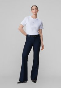 Bílé tričko s krátkými rukávy a malým černým logem, doplněné tmavě modrými rozšířenými kalhotami a černými lodičkami s ostrou špičkou. Jednobarevné, minimalistický design.