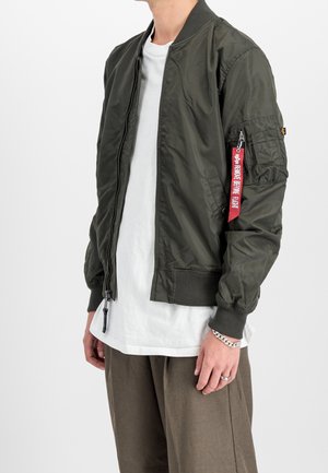 Mężczyzna noszący oliwkowo-zieloną kurtkę bomber z kieszenią na zamek i czerwoną metką "Remove Before Flight" na rękawie, białą koszulę i brązowe spodnie.