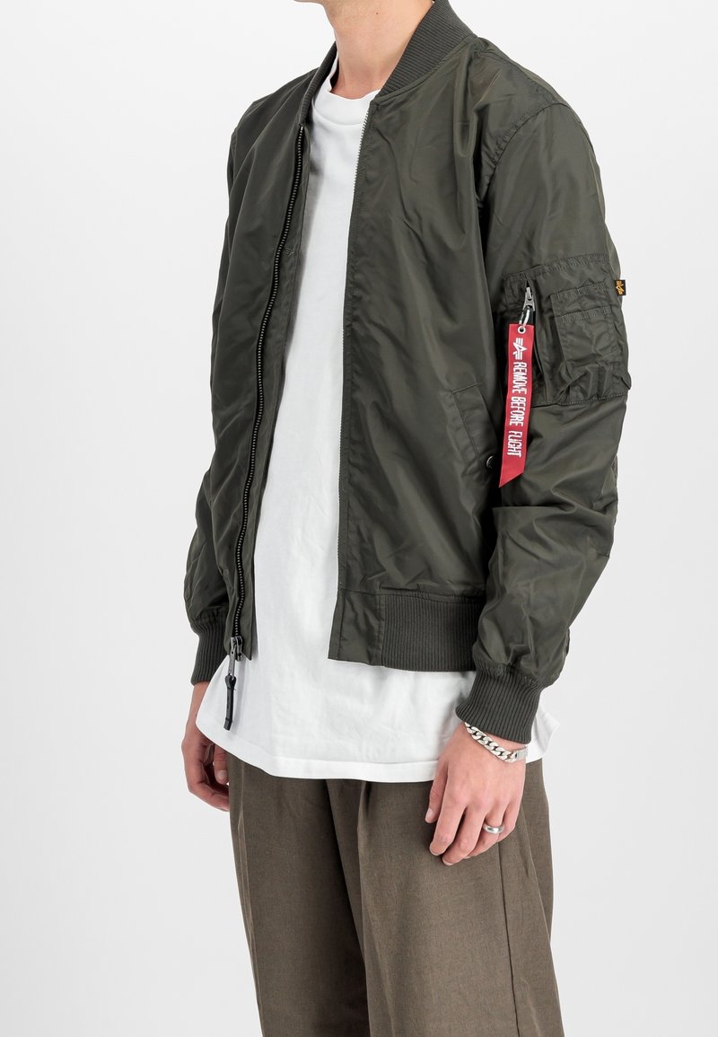 Homme portant une veste bomber vert olive avec une poche zippée et une étiquette rouge « Remove Before Flight » sur la manche, chemise blanche et pantalon marron.