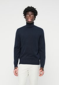 Mornarsko pulover s turtleneck iz mehkega materiala, ki se ponaša z oprijetim silhuetom, rebrastimi manšetami in robom. Usklajen z svetlimi hlačami.