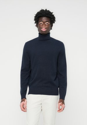 Navy turtleneck sweater in soft material, featuring a fitted silhouette, ribbed cuffs, and hem. Paired with light-colored pants.