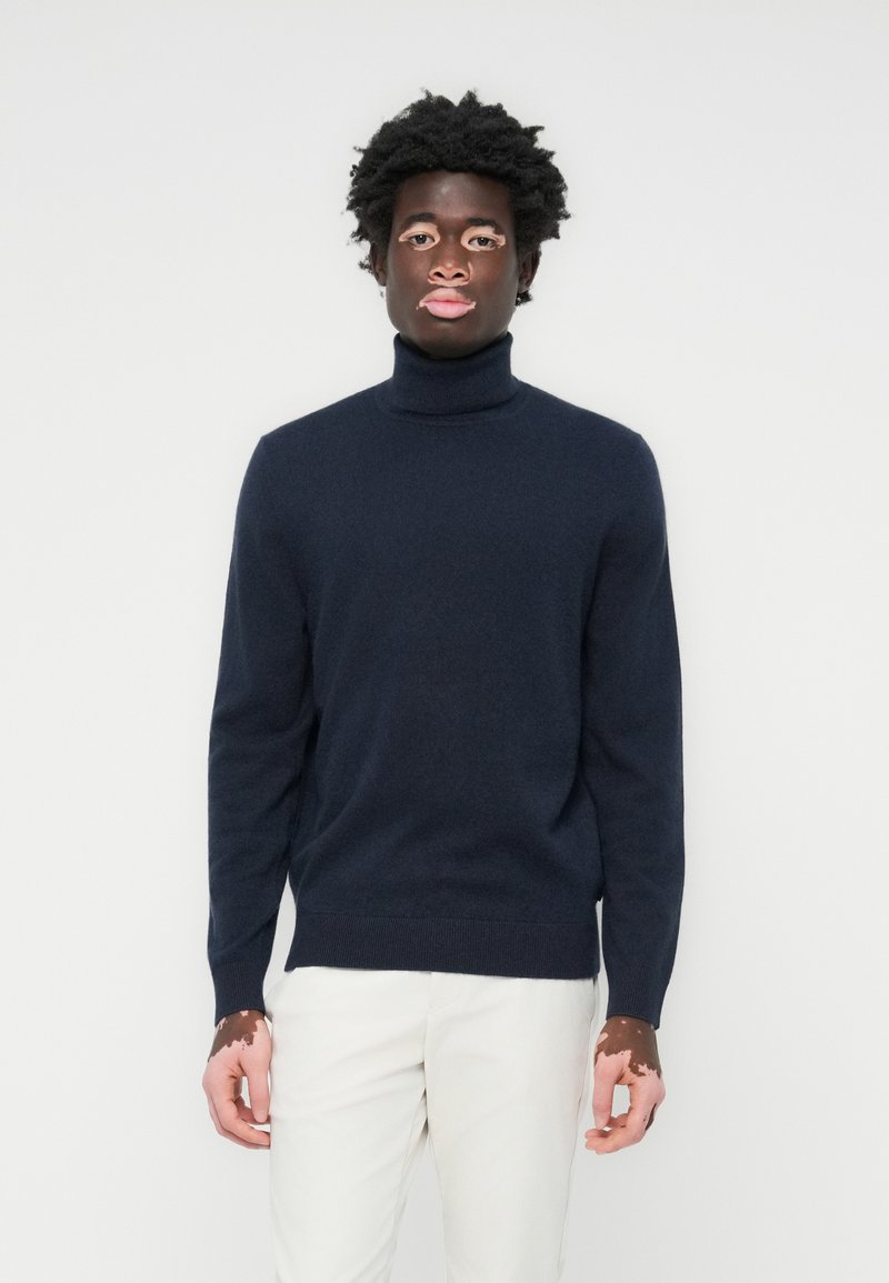 Mornarsko pulover s turtleneck iz mehkega materiala, ki se ponaša z oprijetim silhuetom, rebrastimi manšetami in robom. Usklajen z svetlimi hlačami.