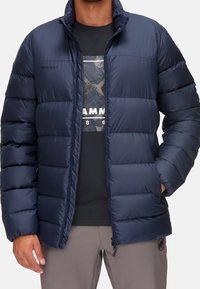 Giacca in nylon blu navy imbottita, con sezioni trapuntate, colletto alto e zip frontale, abbinata a una maglietta scura sotto.