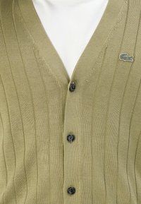 Cardigã canelado em verde oliva claro com botões pretos, por cima de uma camisa branca, apresentando um pequeno logo de crocodilo em verde escuro no peito.