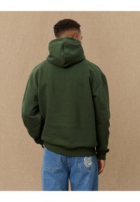 Green hoodie with a large hood, relaxed fit, and ribbed cuffs. Worn with blue jeans featuring a patterned back pocket detail.