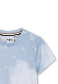 Chemise à manches courtes bleu clair ornée de motifs de logo blancs, avec un col rond à pressions et un col intérieur rayé de couleur contrastée.