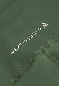 Maslinasto zeleni materijal s glatkom teksturom, s bijelim logotipom "NEXT.STUDIO" i trokutastim akcentom. Vidljive šavove ističu dizajn.