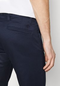 Chino in cotone blu navy con vestibilità slim. Presenta una tasca posteriore e passanti per cintura. Dettagli di cucitura a vista lungo le cuciture.