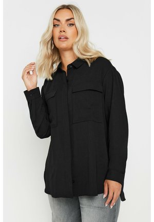 Chemise noire à manches longues avec col, deux poches poitrine, devant à boutons, fabriquée en tissu léger. Le modèle pose avec la main levée.