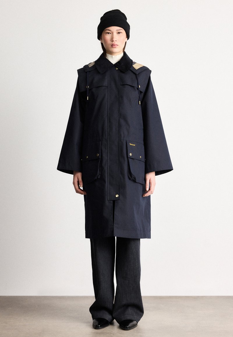 Ανθεκτική στο νερό παρκά σε ναυτικό μπλε με μεγάλες μπροστινές τσέπες, χρυσαφένιες λεπτομέρειες και επισημάνσεις στους ώμους, συνδυασμένη με μαύρο παντελόνι φαρδιάς γραμμής.