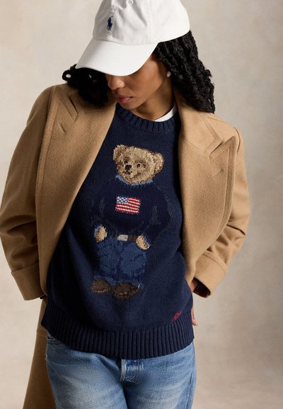 Moteris dėvi smėlio spalvos paltą, tamsiai mėlyną megztinį su meškiuku, dėvinčiu megztinį su Amerikos vėliava, šviesiai mėlynas džinsas ir baltą kepurę.