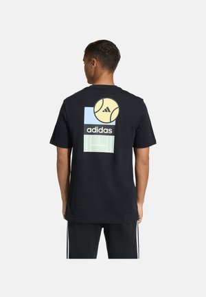 Homme portant un t-shirt noir Adidas avec une balle de tennis, le logo Adidas et un graphique de terrain au dos, associé à un pantalon noir à rayures blanches.