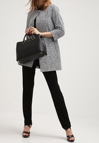 Femme portant un pantalon noir et une veste à carreaux, tenant un grand sac noir, se tenant les jambes écartées sur un fond clair.