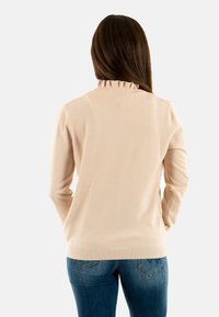 Lichtbeige trui met een gestructureerd stippenpatroon, lange mouwen en een ruches rond de hals. Draag met blauwe jeans.