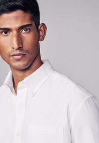 Camicia bianca con colletto, dotata di una tasca sul lato sinistro e realizzata in un morbido cotone, abbinata a uno sfondo neutro.