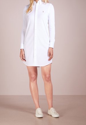Vrouw die een witte jurk met lange mouwen en knopen draagt, met kraag en witte sneakers, staand op een effen beige vloer en achtergrond.