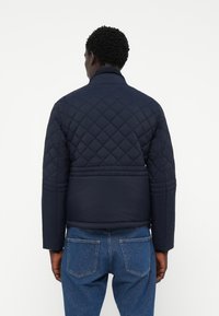Veste matelassée bleu marine avec un col structuré, des manches droites et une longueur cropped. Le tissu semble lisse avec des coutures verticales.