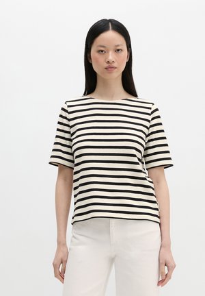 Frau mit langen schwarzen Haaren, die ein kurzärmeliges schwarz-weiß horizontal gestreiftes Shirt und weiße Hosen trägt, steht vor einem schlichten Hintergrund.