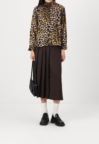 Braun-schwarz gepunktete Bluse mit langen Ärmeln, kombiniert mit einem dunklen plissierten Rock. Eine schwarze Tasche und klobige Slipper runden das Outfit ab.