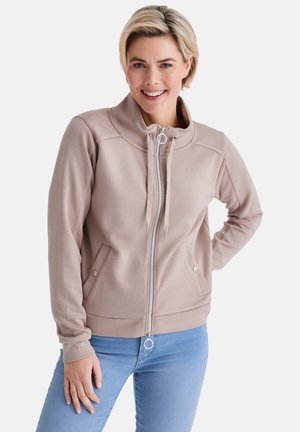 Lächelnde Frau mit kurzen blonden Haaren, die eine beige Jacke mit Reißverschluss und hellblaue Jeans vor weißem Hintergrund trägt.