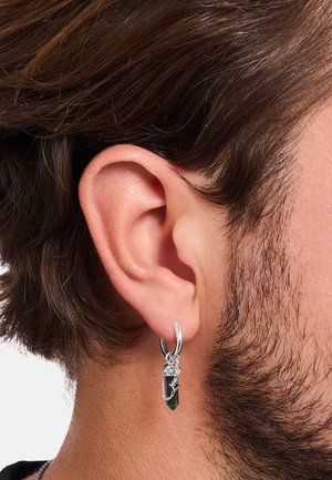 Close-up of a silver hoop earring with a black crystal pendant and decorative chains worn on a man's left ear with brown hair and stubble.