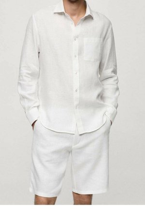 Homme portant une chemise blanche à manches longues avec poche poitrine et un short blanc assorti jusqu'aux genoux, les mains dans les poches, sur un fond uni.