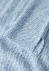 Jasnoniebieska, teksturowana wełniana tkanina z widocznym mankietem rękawa, ukazująca delikatny wzór dzianiny i miękką, puszystą powierzchnię.