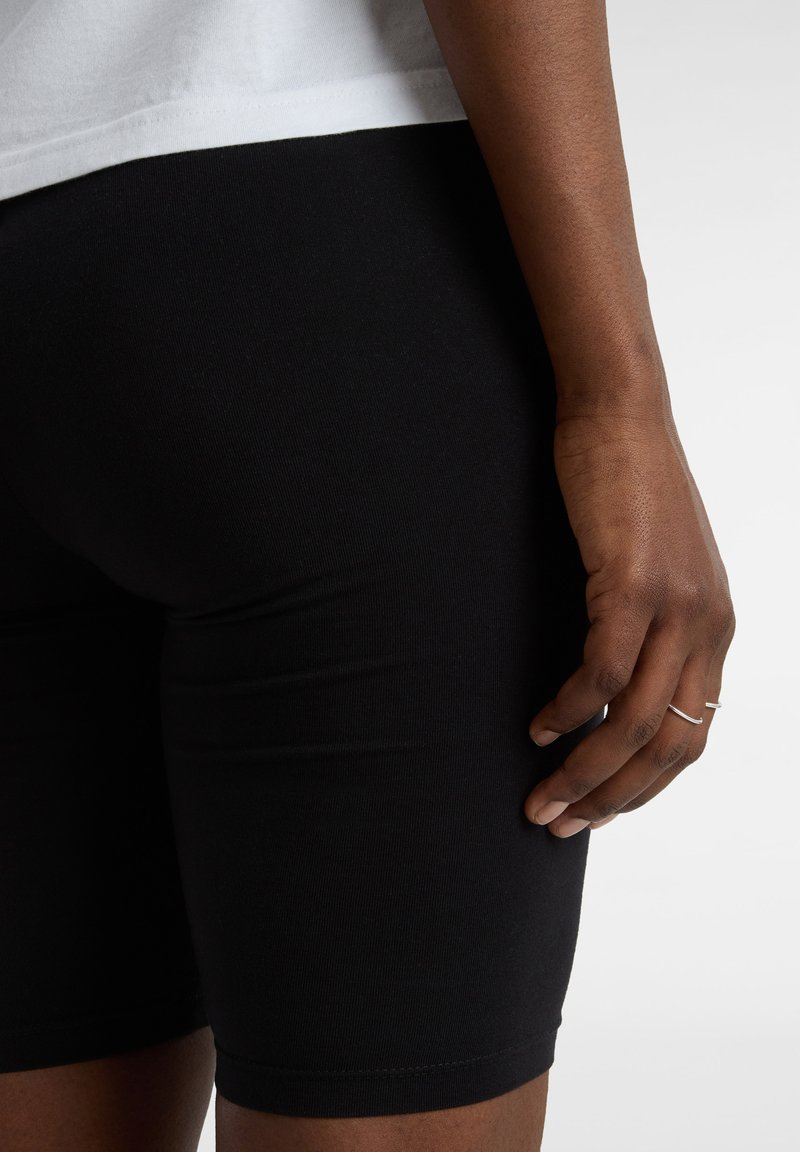 Pantalones cortos ajustados negros de tela elástica, combinados con una blusa blanca. La mano que descansa sobre la pierna muestra un anillo de plata delgada.