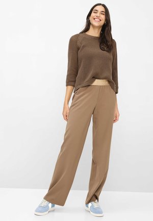 Donna sorridente in piedi che indossa un maglione marrone lavorato a maglia, pantaloni beige larghi e sneakers blu e bianche, su uno sfondo bianco uniforme.