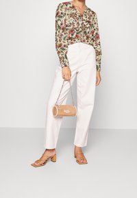 Bluse mit floralem Muster in cremigem Beige und bunten Prints. Weiße, hochtaillierte Hose, beige zylindrische Handtasche mit silberner Kette, Sandalen mit Blockabsatz und Riemchen.