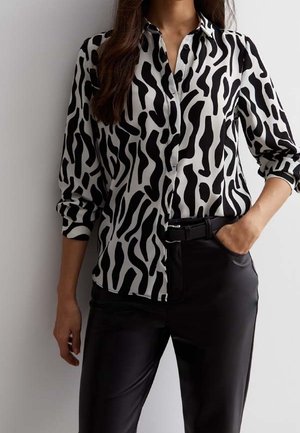 Donna che indossa una camicia con motivo astratto bianco e nero, con maniche arrotolate, e pantaloni neri con una cintura.