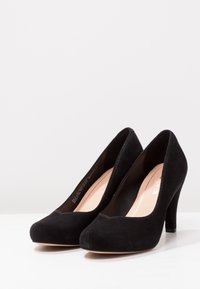 Chaussures à talons hauts en suede noir avec un bout rond et un design sans couture. Elles présentent un intérieur lisse et une semelle extérieure beige.