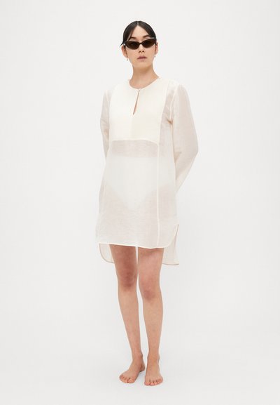 Vestito color beige chiaro a maniche lunghe con scollo a goccia, tessuto trasparente e spacchi laterali, abbinato a occhiali da sole scuri. Design minimalista.