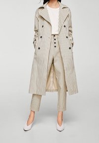Cappotto trench a righe in crema e beige con bottoni doppi, vita abbottonata e pantaloni a righe coordinati. Abbinato a scarpe bianche.
