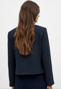 Tamsiai mėlynas tekstūrinis švarkas su trumpu dizainu ir ilgomis rankovėmis. Nugaros vaizdas rodo lygaus paviršiaus apdailą be matomų užtrauktukų ar akcentų.