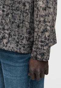 Δεξί χέρι ατόμου που φοράει μακρυμάνικο πουκάμισο με μπεζ και μαύρο σχέδιο, με μπλε τζιν τζιν παντελόνι να φαίνεται κάτω από το τελείωμα του πουκάμισου.